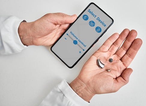 Doctor audiologist showing smartphone app for adjusting hearing aid holding smartphone in one hand and BTE hearing aid in other. Manage hearing aid settings via smartphone