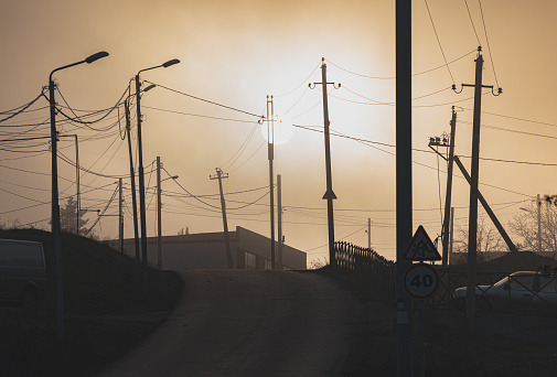 Setting sun in haze hanging over a suburb road with power poles and street lights.