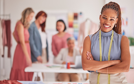 Seamstress, fashion and creative with a black woman designer standing arms crossed in her office with colleagues. Portrait, workshop or measuring tape with a female design employee and team at work