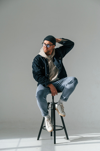 Model in hat and glasses is sitting on a chair. Handsome man is in the studio against white background.