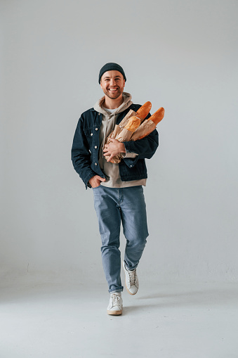 Guy is walking and holding baguettes. Handsome man is in the studio against white background.