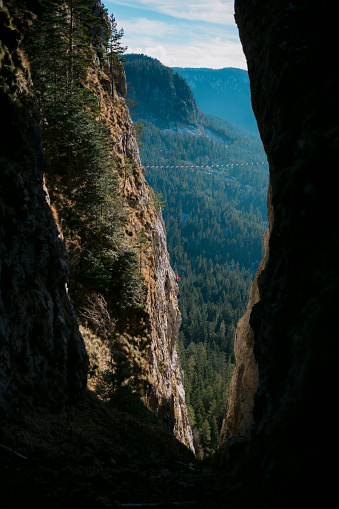 View of a suspension bridge over a deep canyon with mountaineers climbing rock face. Rope bridge in the mountains over the abyss against the backdrop of green valley.