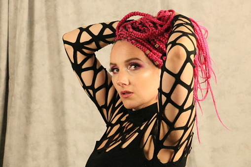 A Caucasian woman playing with her long braided pink hair. She is wearing a black dress and makeup.