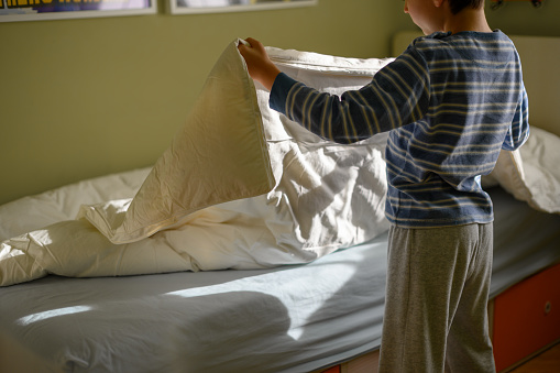 Boy stretches the quilt to make his bed in his room.