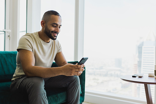 Bearded man sitting on couch in modern apartment, smiling at cellphone