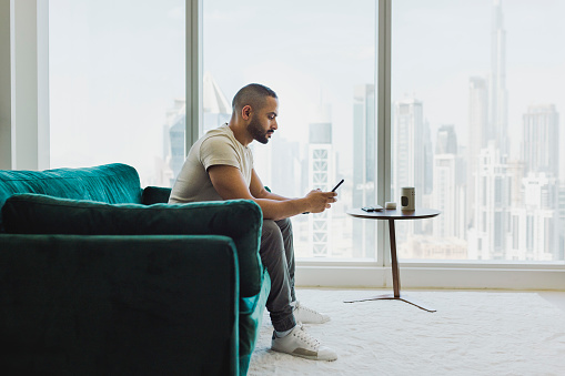 Bearded man sitting on couch in modern apartment with city view, texting on cellphone