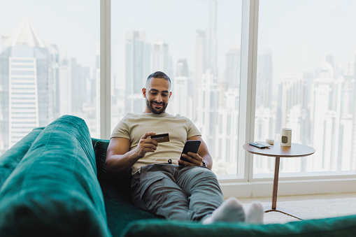 Cheerful bearded man relaxing on couch, shopping with credit card and cellphone in modern apartment with city view