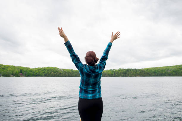 Feeling Limitless Young woman with her back to the camera, faces a beautiful, clear lake with her arms up with joy and freedom. She is wearing a blue, plaid shirt, and there is only sky, trees and water ahead of her. cottage life stock pictures, royalty-free photos & images