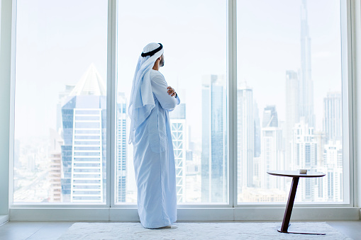 Arab businessman wearing kandora, looking through window in modern office with city view