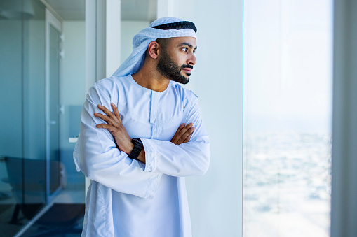 Bearded Arab man wearing kandora and crossing arms looking through window in office