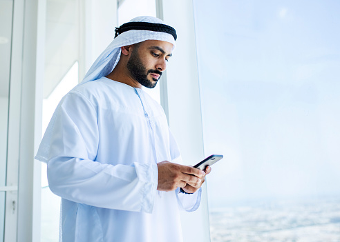 Hombre árabe usando un teléfono celular junto a la ventana de la oficina photo