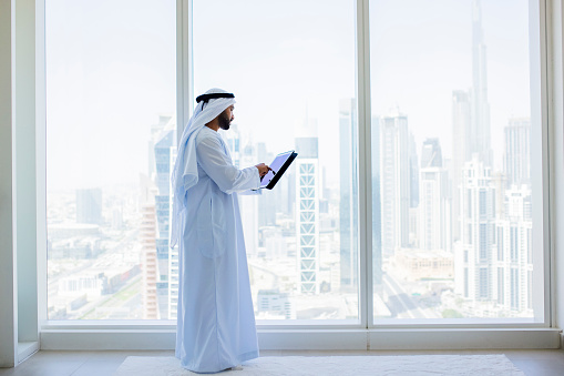 Bearded Arab businessman wearing kandora, working on digital tablet beside window in modern office with city view