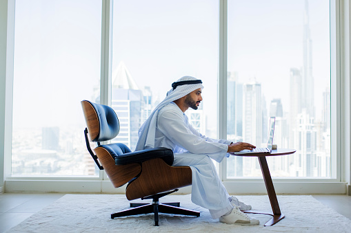 Bearded Arab businessman sitting on modern armchair in office with city view, concentrating with work on laptop