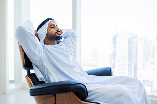 Arab man with beard, closing eyes and relaxing with hands behind head on armchair in modern office with city view