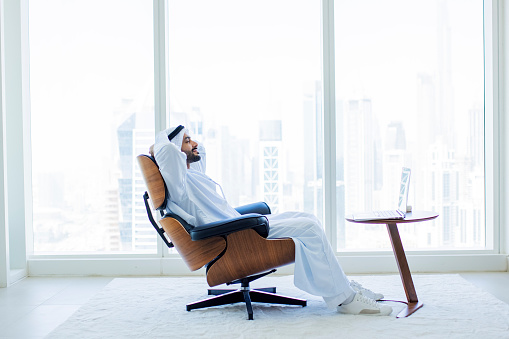 Happy Arab man with beard, resting with eyes closed and hands behind head on armchair in modern office with city view