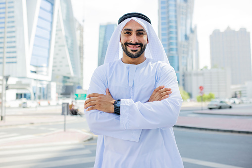 Bearded Arab businessman wearing kandora, crossing arms, smiling and looking at camera on the streets in city centre