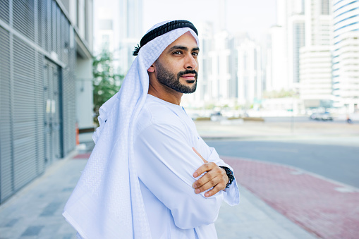 Bearded Arab businessman wearing kandora, crossing arms, standing on pavement and looking into distance in city centre