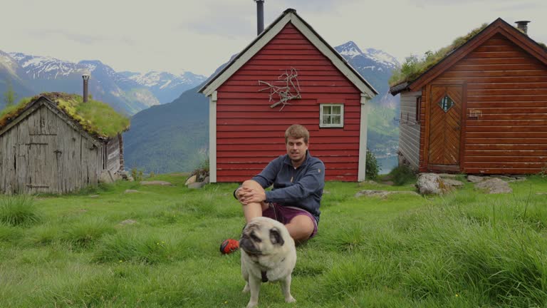 Front view of a happy man with dog contemplating the scenic mountain valley with old wooden houses in Norway