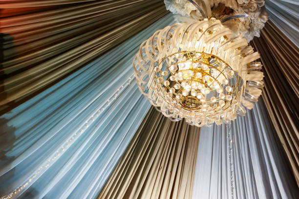 Low angle close up view of modern chandelier hanging from ceiling with decorative white and brown curtain sheets. No people.