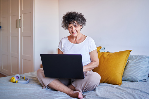 Front view of a relaxed mature woman surfing the net with her laptop sitting on bed.
