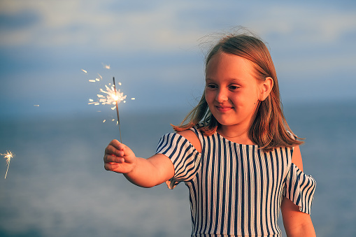 happy girl of eight years old with sparklers in her hand and in a dress with blue stripes on the ocean shore on a holiday