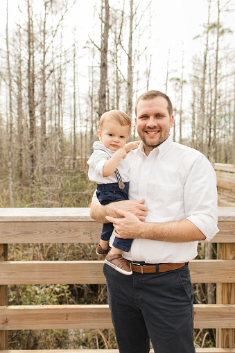 A 31-Year-Old American Father Holding His 12-Month-Old Cuban-American Son Dressed in Classic White & Navy on a Boardwalk in a Woodsy Forest
