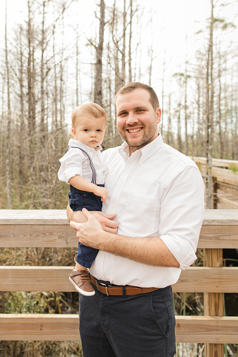 A 31-Year-Old American Father Holding His 12-Month-Old Cuban-American Son Dressed in Classic White & Navy on a Boardwalk in a Woodsy Forest