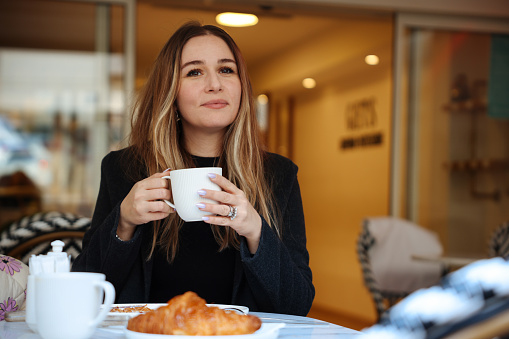 Happy young woman having cafe latte and croissant in cafe