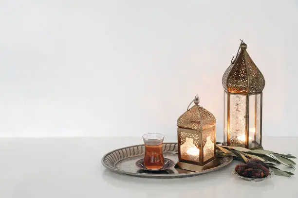 Festive Ramadan Kareem still life. Glowing Moroccan ornamental lanterns and glass cup with Turkish tea. Olive tree branches, date fruit on silver tray, white wall background. Empty copy space, indoor.