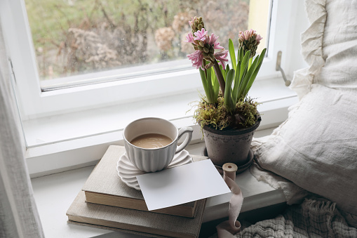 Feminine spring composition. Cup of coffee, books and blank greeting card mockup near window. Vintage Easter styled photo. Moody floral composition. Potted pink hyacinth flowers, linen cushion.