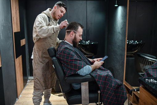 Male Customer Surfing The Net On Smartphone While Getting Professional Haircut Done By Barber