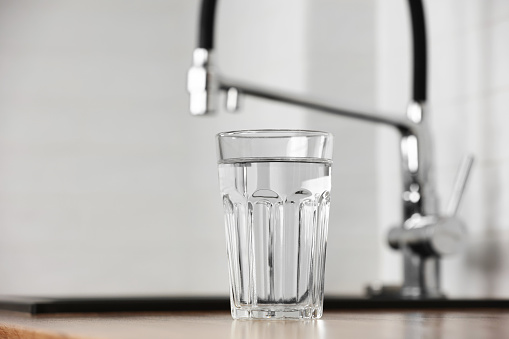 Transparent glass of clean filtering water on wooden table in kitchen interior. Tap with purified water with an osmosis system of the home kitchen sink.