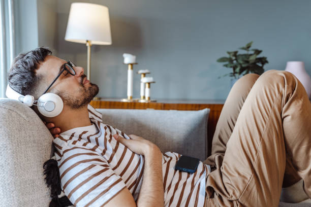 joven usando auriculares inalámbricos y relajándose en el sofá - chill out audio fotografías e imágenes de stock