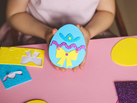 The hands of a little caucasian girl show the end result of a homemade felt easter egg, sitting at a children's table with a set of crafts on a pink background with depth of field, close-up side view. The concept of crafts, diy, needlework, diy, children art, artisanal, Easter preparation, children creative.