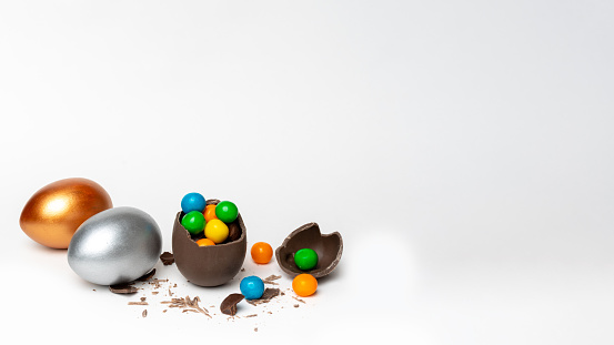 Cracked chocolate easter egg with colorful small round sweets and chocolates and whole colored eggs on white background, copy space. Easter concept.