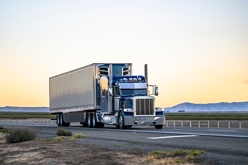 Industrial long hauler classic big rig dark blue semi truck tractor transporting cargo in refrigerator semi trailer driving on the highway road between the field in California at twilight