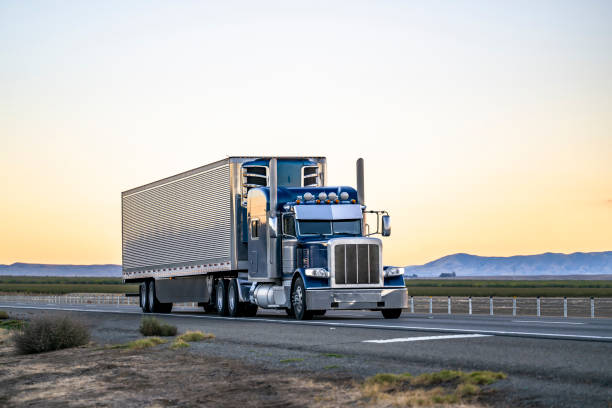 Heavy duty dark blue classic stylish big rig semi truck tractor with lot of chrome accessories transporting cargo in refrigerator semi trailer driving on the road at twilight Industrial long hauler classic big rig dark blue semi truck tractor transporting cargo in refrigerator semi trailer driving on the highway road between the field in California at twilight semi truck stock pictures, royalty-free photos & images