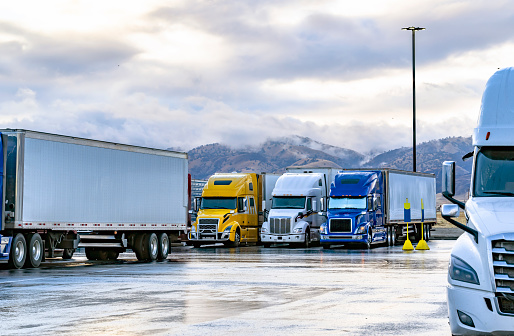General logistic transportation big rigs semi trucks with semi trailers standing in row on truck stop parking lot with wet surface for truck driver rest waiting for the continuation freights