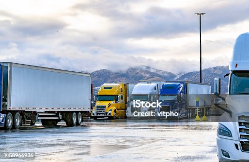 istock Different big rigs semi trucks with loaded semi trailers standing on the truck stop parking lot with wet surface and mountains on the background 1458966936