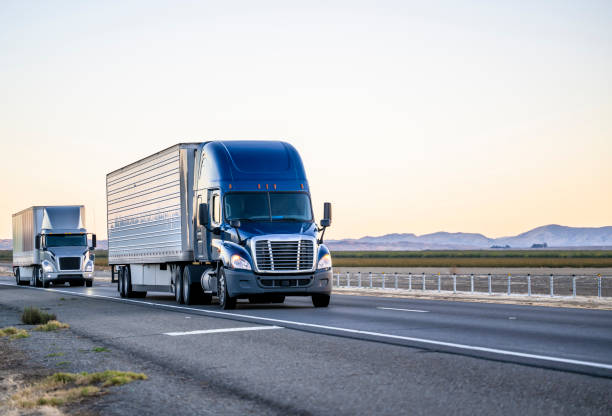 Two industrial bonnet big rigs semi trucks transporting cargo in different semi trailers driving on the straight highway road in California stock photo