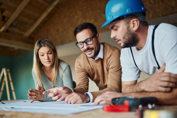Young couple analyzing blueprints with their construction worker/ Young couple and their manual worker cooperating while examining housing plan at construction site. building contractor stock pictures, royalty-free photos & images