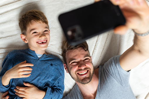 Young father and son having fun in bedroom, making selfie. High angle view.