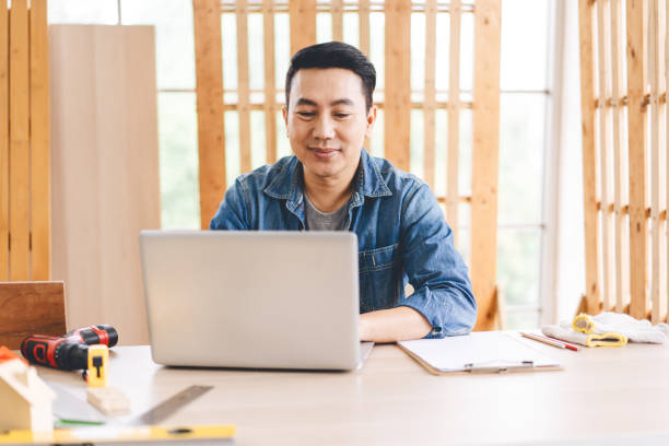 mittelalter erwachsener asiatischer mann tischler mit laptop für online-arbeit mit kunden wotk zu hause - southeast asian ethnicity men laptop image type stock-fotos und bilder