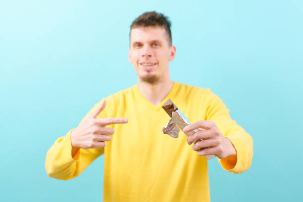 istock A man holds a chocolate bar in a pyramid shape and points a finger at it on the blue background. Taste, One, Orange, Yummy, Nuts, Block, Dairy Product, Design, Triangular, Decorative, Appetizing 1458993782