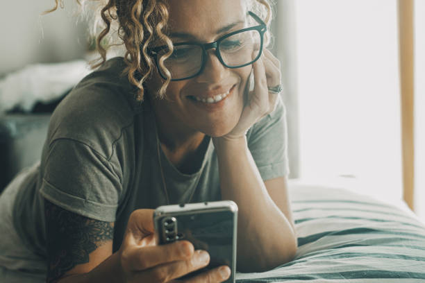 Portrait of cheerful happy woman laying on bed and using mobile phone to chat and watch online contents. Modern female people with smartphone. Chatting with friends. Relax with technology. Writing stock photo