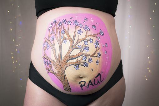 A young pregnant woman who is waiting for every moment of childbirth calmly poses and enjoys while the artist draws her belly. Happy pregnant woman. Body painting is in progress. The art of drawing on the body on the belly of a pregnant woman