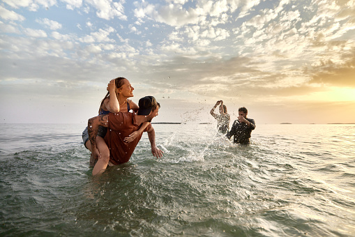Young cheerful couples having fun while splashing during summer vacation in sea at sunset. Focus is on couple piggybacking. Copy space.