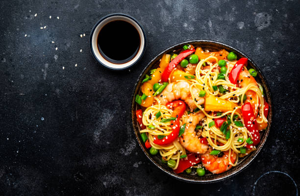 Stir fry noodles with shrimps, red and yellow paprika, green pea, chives and sesame seeds bowl. Asian cuisine dish. Black stone kitchen table background, top view stock photo