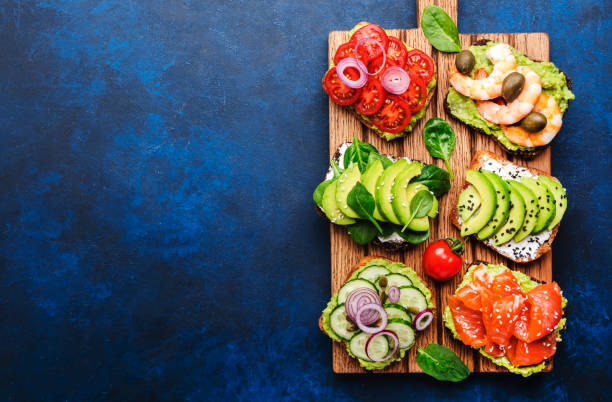 sandwichs à l’avocat ou ensemble de toasts: avec saumon, crevettes, tomates, concombres, fromage à pâte molle et épinards, noix de cajou et graines de sésame servis sur planche à découper, fond de table bleu, vue de dessus - appetizer bread breakfast cashew photos et images de collection