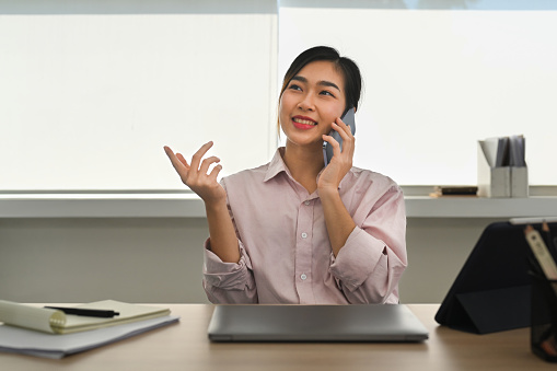 Positive asian female investor having phone conversation with business partners while sitting at her workplace.
